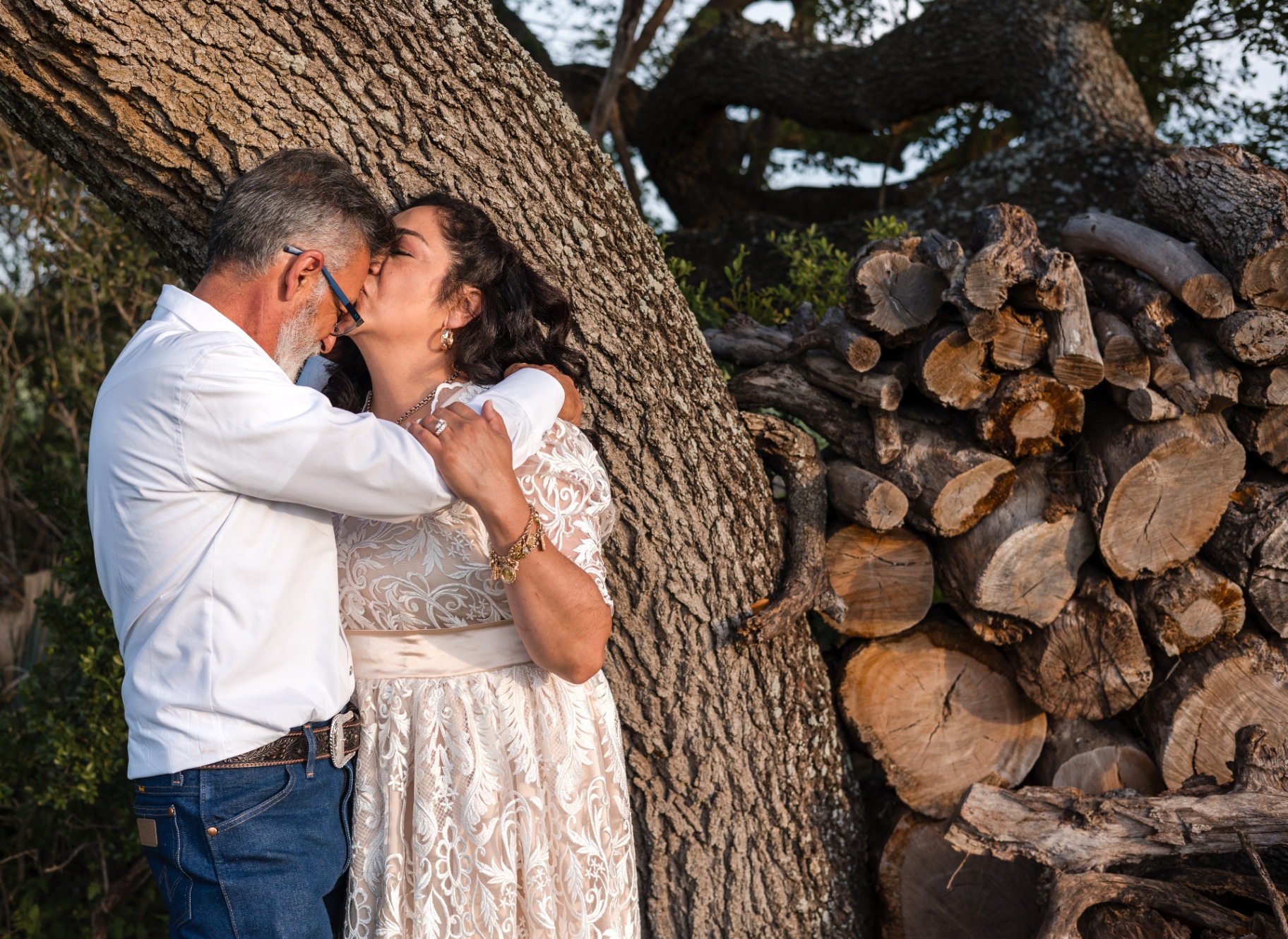 Couple sharing a kiss under a large oak tree beside stacked firewood