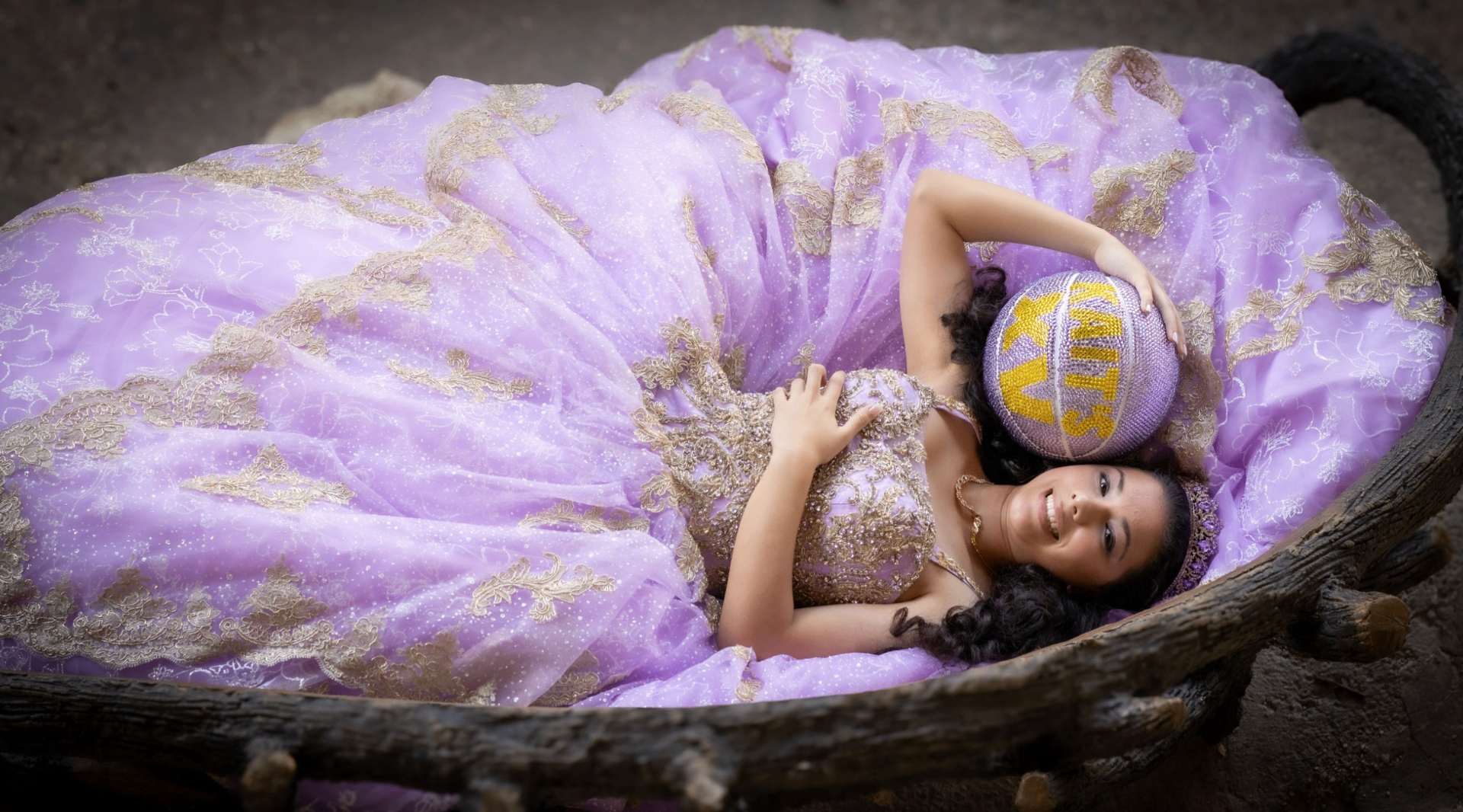 Quinceañera in lilac gown lying in a wooden cradle holding a XV basketball